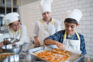 A young kid in plaid shirt making a pizza while standing near his friends